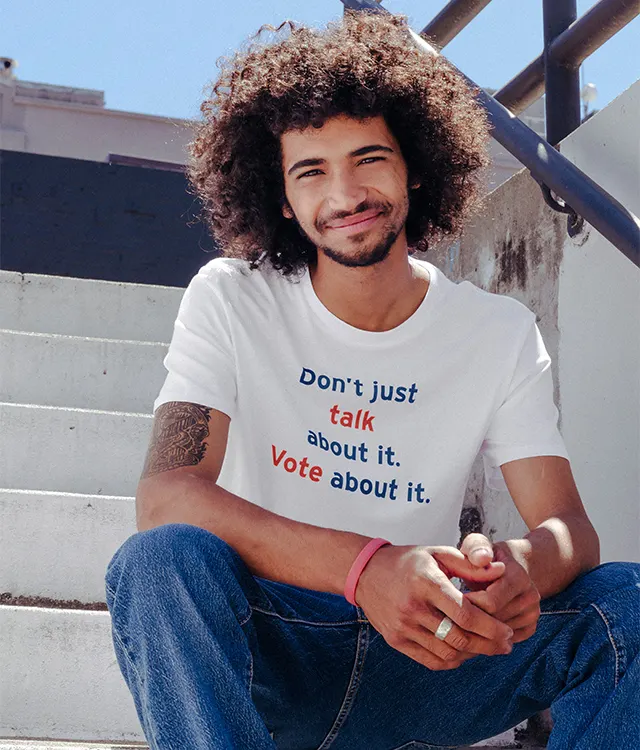 A smiling young man with curly hair sits on outdoor steps, wearing a white shirt that says “Don’t just talk about it. Vote about it.” in blue, orange, and red letters.