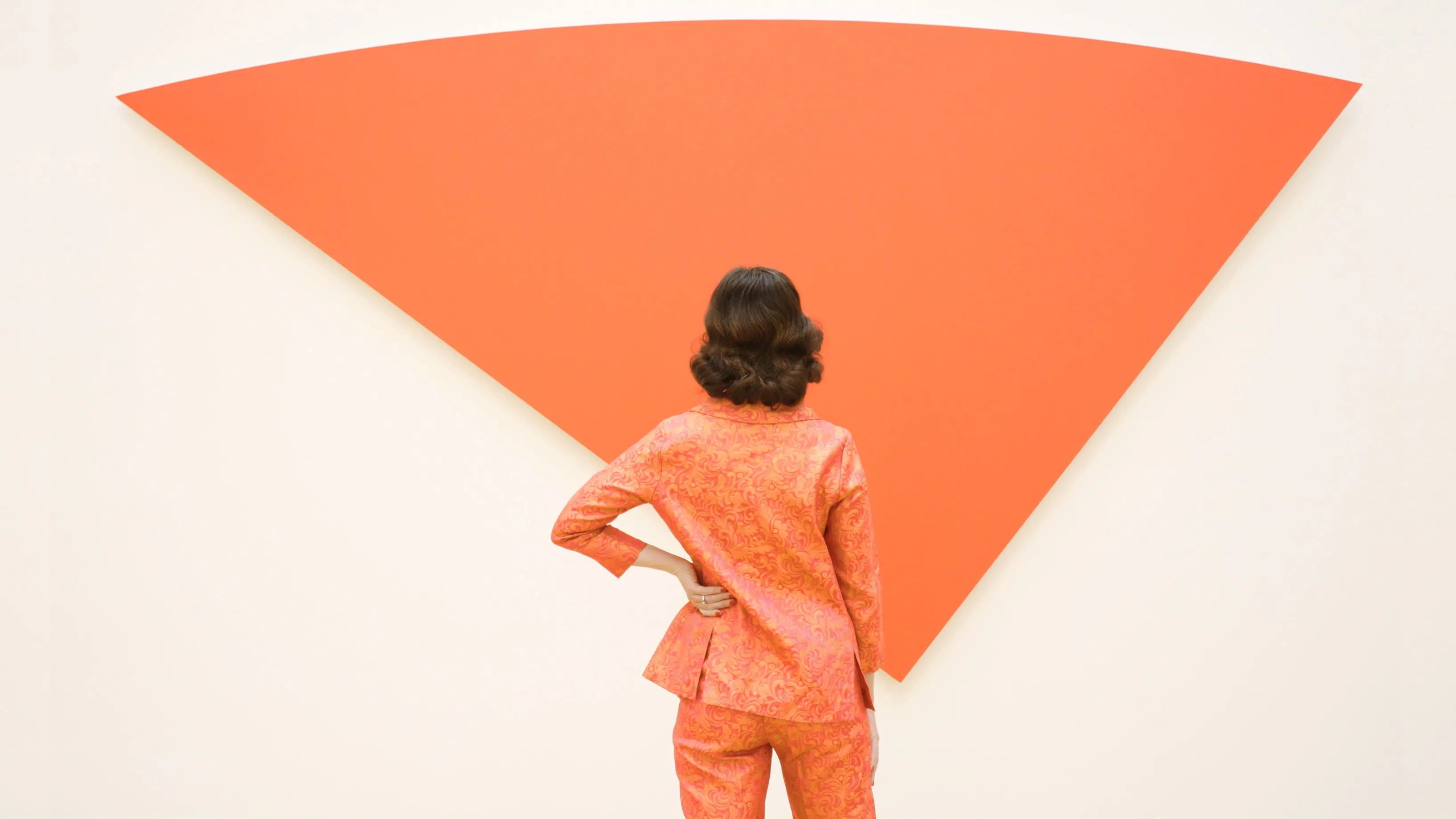 A person in a patterned orange suit stands facing a large, inverted red triangle art piece mounted on a white wall.