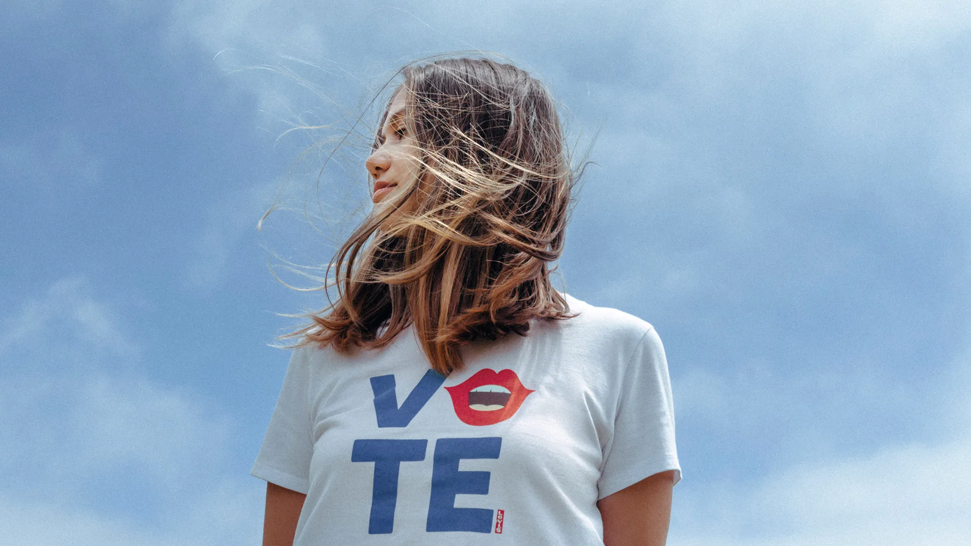 A person with shoulder-length hair stands against a blue sky, wearing a white T-shirt that says VOTE with a red lips graphic in place of the letter O. The person looks to the side.